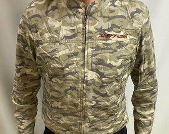 May include: A camouflage-patterned jacket with a stand-up collar and a full-length zip. The jacket features the Harley-Davidson logo in orange on the chest. The person in the image is wearing black trousers and boots.