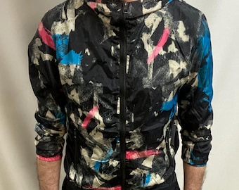 May include: A black hooded jacket with a full-length zip, adorned with a vibrant abstract design in blue, pink, and white. The jacket is worn with black trousers. The ensemble presents a casual, sporty aesthetic.