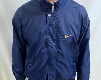 May include: Navy blue Nike windbreaker jacket with a yellow swoosh logo. The jacket features a stand-up collar, zip closure, and two side pockets. The jacket is worn with black trousers.