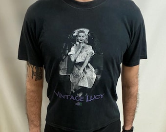 May include: Black crew neck t-shirt with a black and white image of a woman and the text "VINTAGE LUCY" in purple. The t-shirt is a classic style.