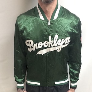 May include: Green satin bomber jacket with the word "Brooklyn" in white script across the chest. The jacket has white and green striped trim on the collar, cuffs, and hem. The model is wearing black pants and black sneakers.