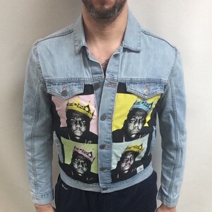 May include: Light blue denim jacket featuring a colorful graphic of a person wearing a crown. The jacket has a classic collar, button-up front, and long sleeves. The person is also wearing navy blue shorts and black high-top sneakers.