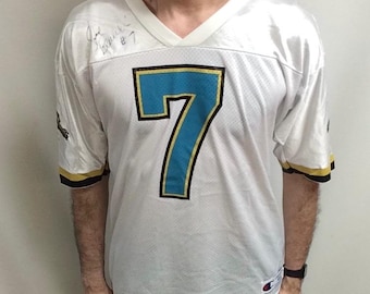 May include: White mesh jersey with the number 7 in teal and black, gold trim on the sleeves and collar. The jersey has a V-neck and a Champion logo. Black shorts and black shoes complete the outfit.