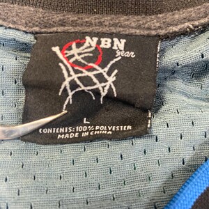 Puede incluir: Primer plano de una etiqueta negra con el texto "NBN gear" y un gr&aacute;fico de red de baloncesto. La etiqueta est&aacute; adherida a una camiseta de malla azul y negra. La etiqueta tambi&eacute;n dice "L" y "CONTENTS: 100% POLYESTER MADE IN CHINA."