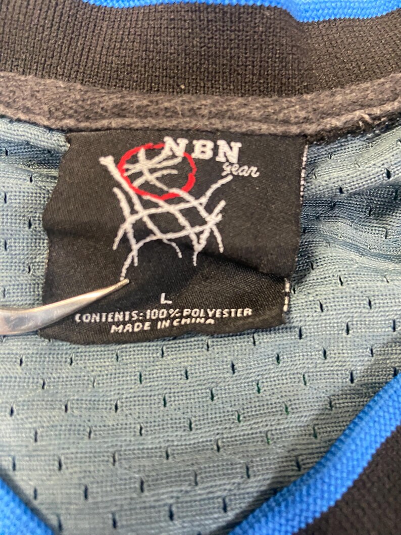 Puede incluir: Primer plano de una etiqueta negra con el texto "NBN gear" y un gr&aacute;fico de red de baloncesto. La etiqueta est&aacute; adherida a una camiseta de malla azul y negra. La etiqueta tambi&eacute;n dice "L" y "CONTENTS: 100% POLYESTER MADE IN CHINA."