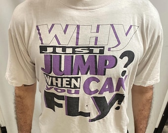 May include: Cream-coloured t-shirt featuring the phrase "WHY JUST JUMP WHEN YOU CAN FLY?" in purple and black. The shirt has a crew neck and short sleeves. The text is in a bold, stylised font.