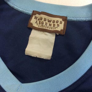 May include: Navy blue athletic jersey with a light blue collar and trim. A brown tag with the words "HARDWOOD LEGENDS" is sewn into the jersey. The tag also includes the text "BY HEADMASTER CAMPUSWEAR".