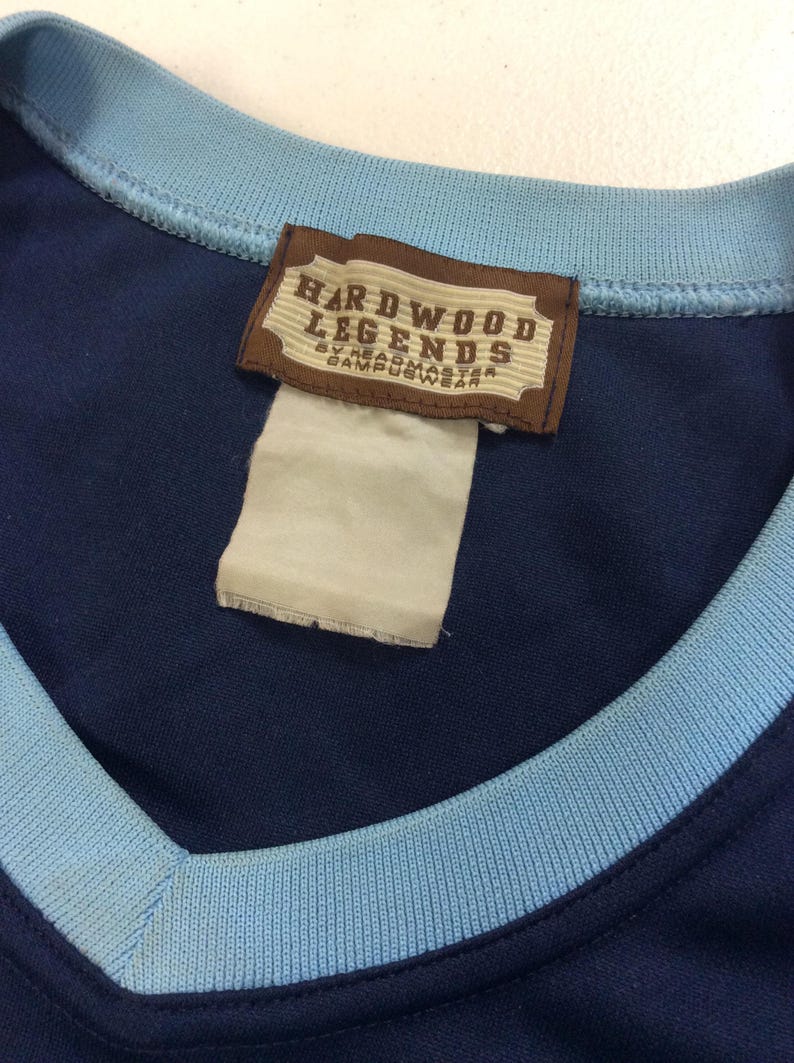 May include: Navy blue athletic jersey with a light blue collar and trim. A brown tag with the words "HARDWOOD LEGENDS" is sewn into the jersey. The tag also includes the text "BY HEADMASTER CAMPUSWEAR".