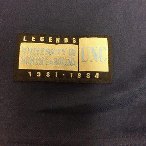 May include: Navy blue fabric with a rectangular patch. The patch has the word "LEGENDS" at the top, with "UNIVERSITY OF NORTH CAROLINA" and "UNC" in light blue. The years "1981-1984" are at the bottom.