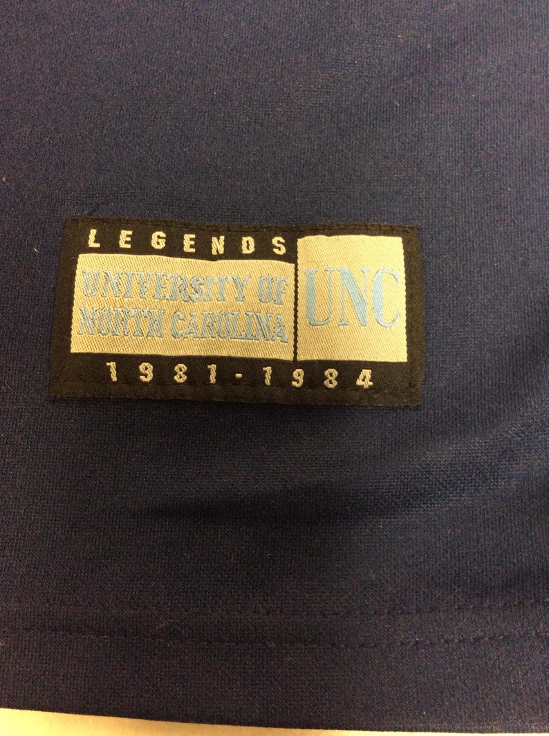 May include: Navy blue fabric with a rectangular patch. The patch has the word "LEGENDS" at the top, with "UNIVERSITY OF NORTH CAROLINA" and "UNC" in light blue. The years "1981-1984" are at the bottom.