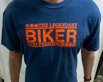 May include: A navy blue t-shirt featuring the orange text "THE LEGENDARY BIKER HARLEY-DAVIDSON". The shirt has a crew neck and short sleeves. The text is in a bold, blocky font, typical of motorcycle apparel.