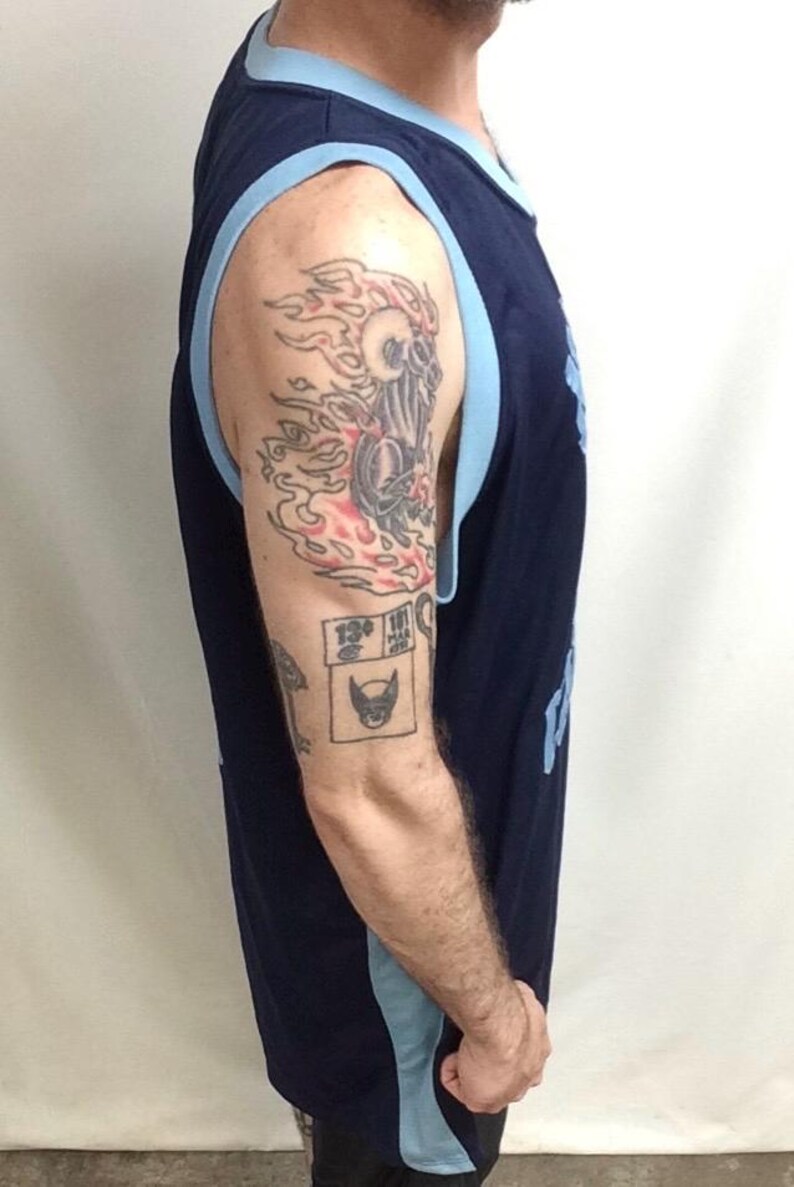 May include: Navy blue sleeveless jersey with light blue trim. The jersey is visible from the side. The arm has tattoos, including a skull with flames and a Wolverine logo.