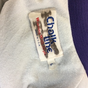 May include: A white tag with the text "Chalk Line" in black and "Made in U.S.A." in blue. The tag is attached to a purple and white jacket with a size "L" tag.