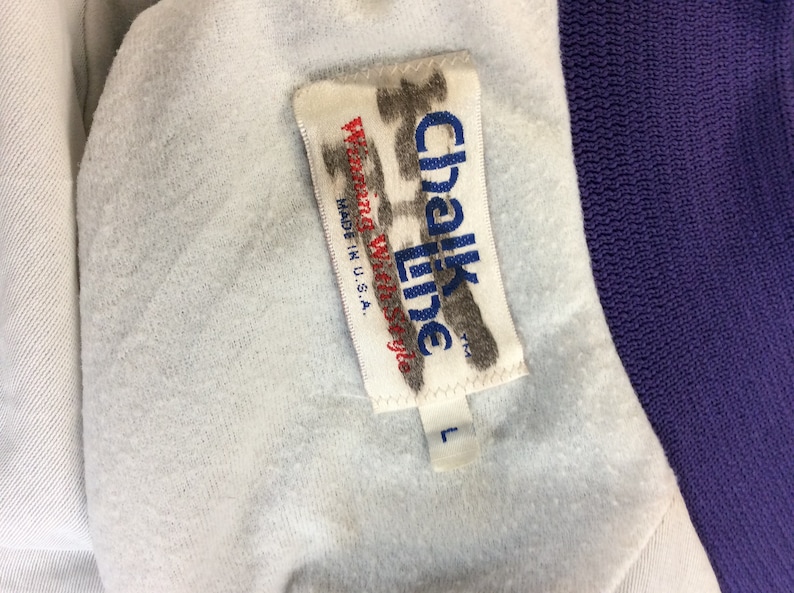 May include: A white tag with the text "Chalk Line" in black and "Made in U.S.A." in blue. The tag is attached to a purple and white jacket with a size "L" tag.