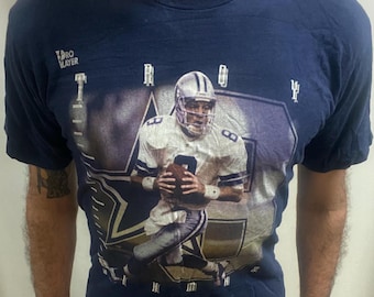 May include: Navy blue t-shirt with a graphic of a football player in a white and blue uniform, holding a football. The shirt features the text "Pro Player" and the Dallas Cowboys logo. A vintage design.