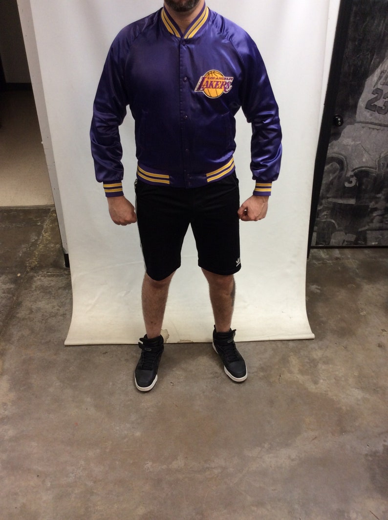 May include: A purple satin bomber jacket with yellow and gold stripes on the cuffs and waistband. The jacket has a large Los Angeles Lakers logo on the left chest.