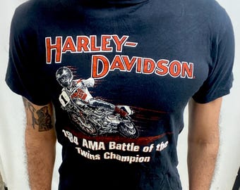 May include: Black Harley-Davidson t-shirt with a red and white logo and a motorcycle racer graphic. The shirt includes the text "1984 AMA Battle of the Twins Champion".