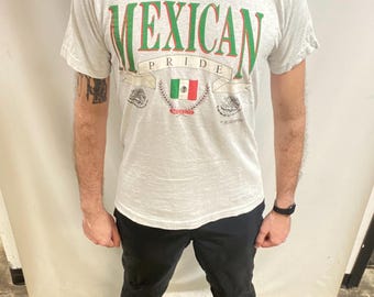 May include: A light grey t-shirt featuring the words "MEXICAN PRIDE" in green and red. Below the text is a banner with the Mexican flag and the word "MEXICO". The shirt is worn with black trousers.