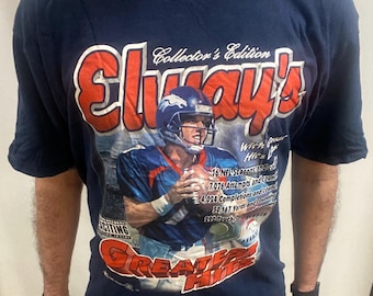 May include: Navy blue t-shirt with a graphic of a football player in blue and orange. The shirt features the text "Elway's Greatest Hits Collector's Edition" and statistics. A vintage sports top.