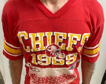 May include: Red vintage Chiefs football jersey with yellow and white detailing. The jersey displays the word "CHIEFS" in large yellow letters, the number "1969", and a football helmet graphic. The sleeves have yellow and white stripes.