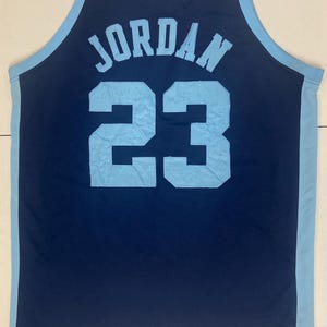 May include: Navy blue basketball jersey with light blue accents. The jersey features the name "JORDAN" in light blue block letters above the number "23". The number "23" is also in light blue. The jersey is sleeveless.