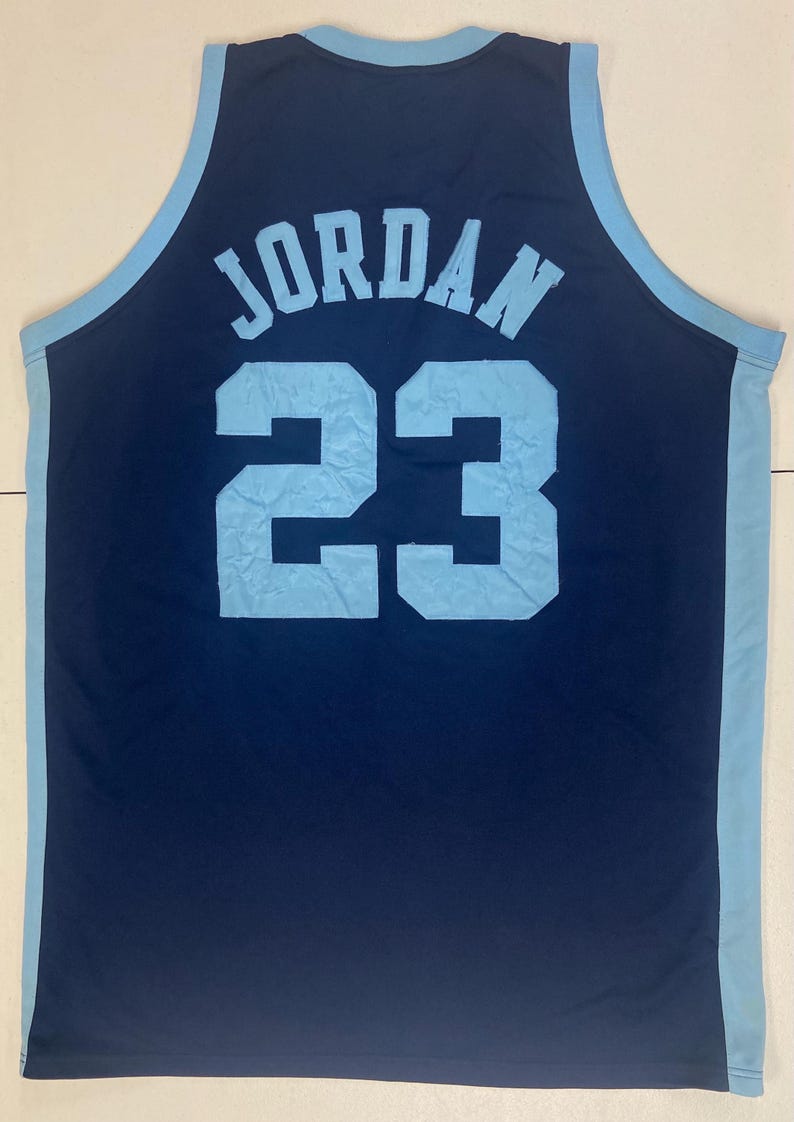 May include: Navy blue basketball jersey with light blue accents. The jersey features the name "JORDAN" in light blue block letters above the number "23". The number "23" is also in light blue. The jersey is sleeveless.