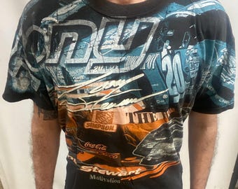 May include: Black t-shirt with a racing design, featuring the number 20 in white and blue. The shirt includes sponsor logos like Coca-Cola and Simpson. The design has orange and white accents.