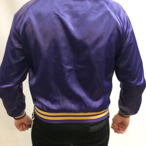 May include: A purple satin bomber jacket with yellow and purple striped trim around the collar, cuffs, and hem.