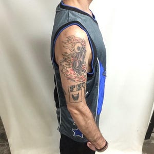 Puede incluir: Camiseta de baloncesto gris y azul con un dise&ntilde;o de estrella azul y blanco. La camiseta tiene un ribete negro. El brazo de la persona tiene un tatuaje de una calavera en llamas y un peque&ntilde;o tatuaje cuadrado con una cara de gato.