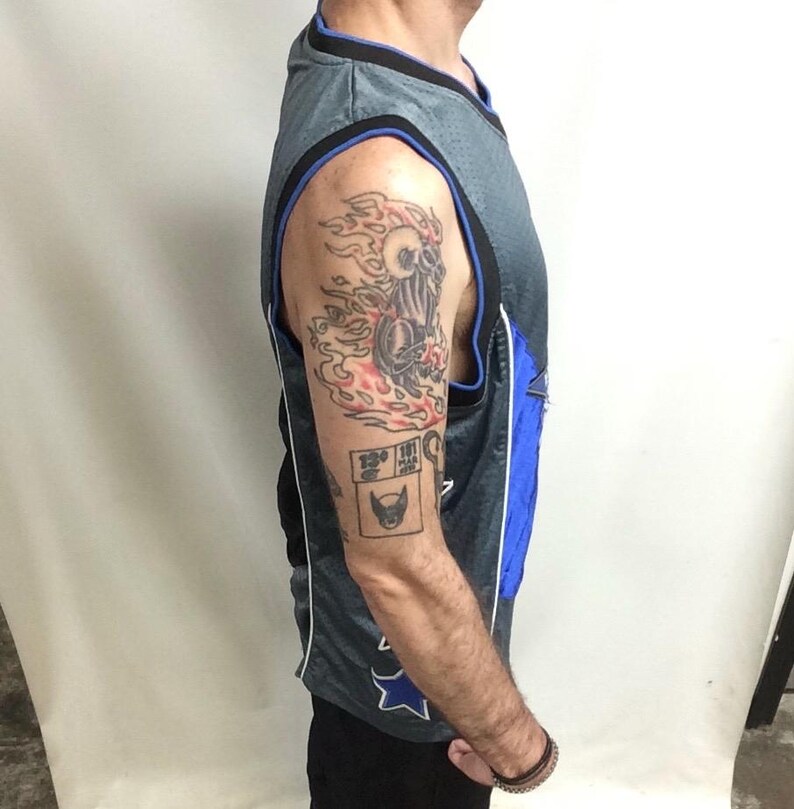 Puede incluir: Camiseta de baloncesto gris y azul con un dise&ntilde;o de estrella azul y blanco. La camiseta tiene un ribete negro. El brazo de la persona tiene un tatuaje de una calavera en llamas y un peque&ntilde;o tatuaje cuadrado con una cara de gato.