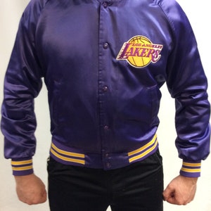 May include: A purple satin bomber jacket with yellow and purple striped cuffs and waistband. The jacket features a large embroidered Los Angeles Lakers logo on the left chest.