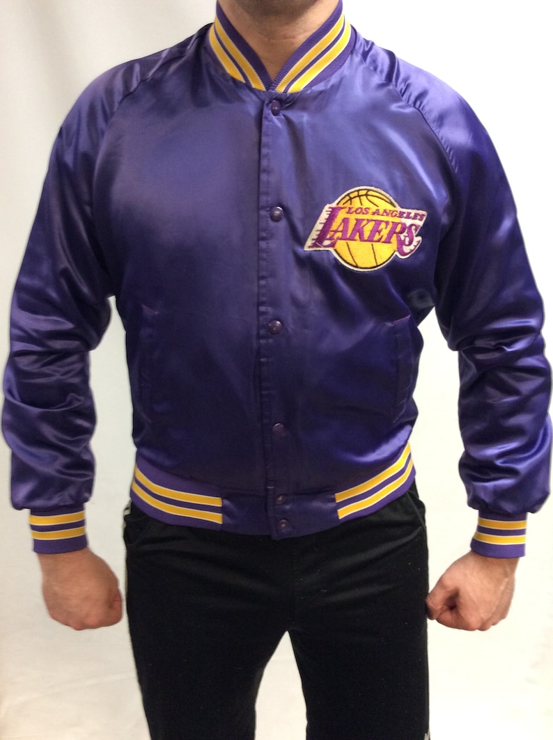 May include: A purple satin bomber jacket with yellow and purple striped cuffs and waistband. The jacket features a large embroidered Los Angeles Lakers logo on the left chest.