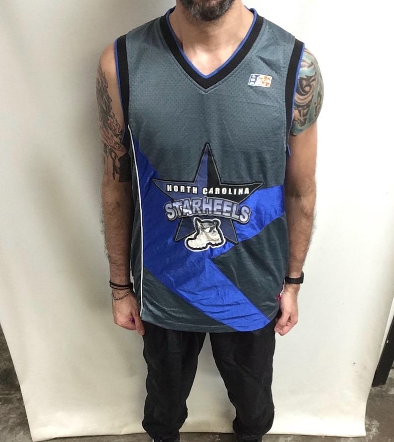 Puede incluir: Camiseta de baloncesto gris y azul con cuello en V y las palabras "North Carolina Starheels" en un dise&ntilde;o de estrella. La camiseta tiene una raya blanca en el lateral y un peque&ntilde;o logotipo en la parte superior derecha.