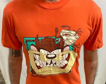 May include: Orange t-shirt with a cartoon image of the Tasmanian Devil and a tornado, with the text "Six Flags". The graphic is set within a teal and yellow frame. The shirt has short sleeves.