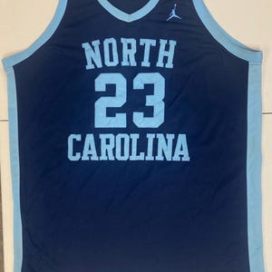 May include: Navy blue basketball jersey with light blue trim. The jersey features the words "NORTH CAROLINA" and the number "23" in light blue. A Jumpman logo is visible on the upper right chest.