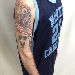May include: Navy blue basketball jersey with light blue trim and the text "NORTH CAROLINA 23 CAROLINA". The jersey is worn by a person with tattoos on their arm, including a flaming ram and a comic book-style image.
