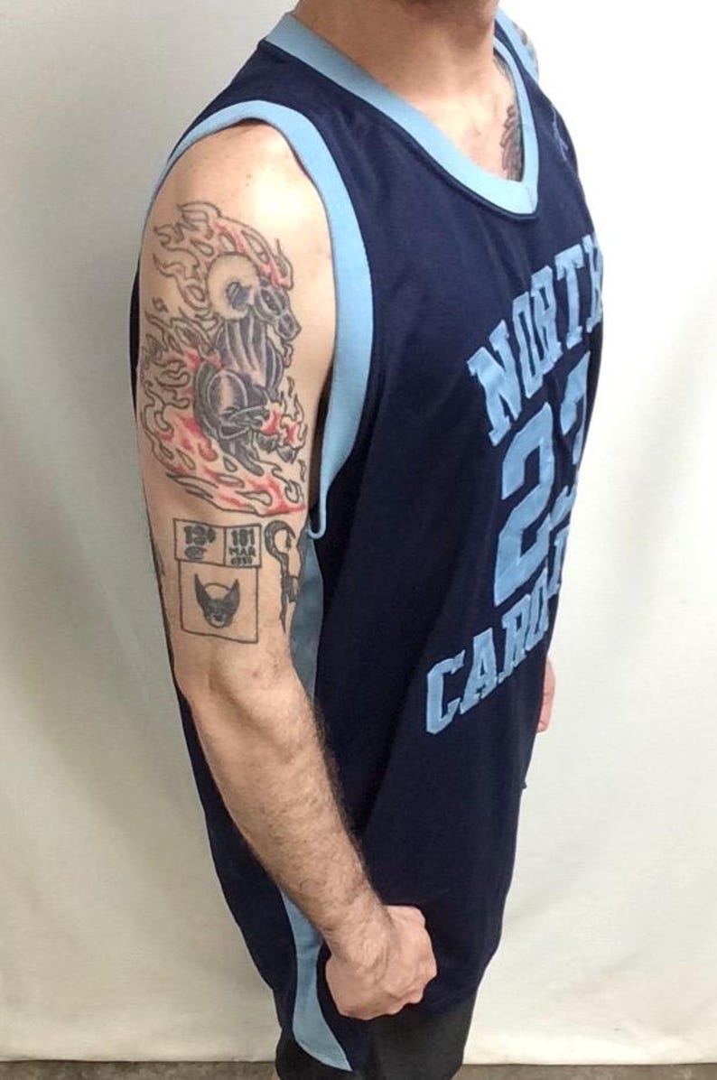 May include: Navy blue basketball jersey with light blue trim and the text "NORTH CAROLINA 23 CAROLINA". The jersey is worn by a person with tattoos on their arm, including a flaming ram and a comic book-style image.