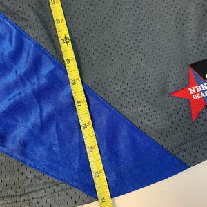 Puede incluir: Tejido de malla gris con una franja diagonal azul. Una cinta m&eacute;trica amarilla muestra una longitud de aproximadamente 79 cm. Una etiqueta roja en forma de estrella con texto blanco es visible en la esquina. La tela parece ser ropa deportiva.