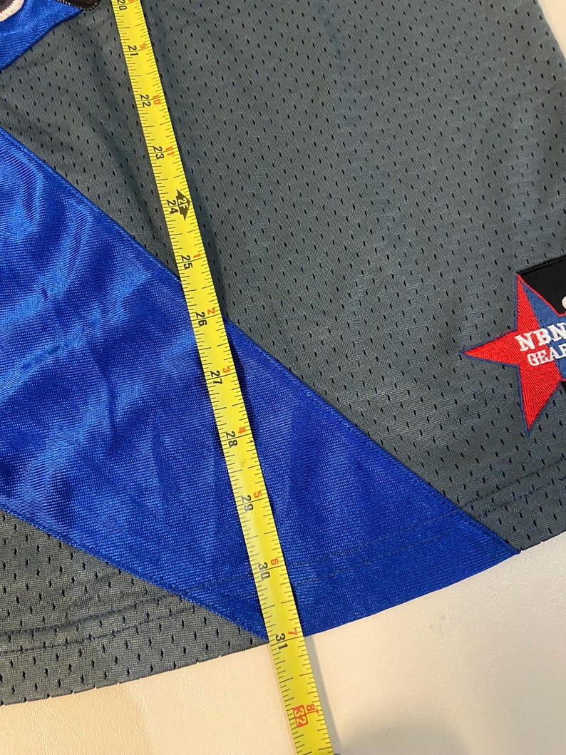 Puede incluir: Tejido de malla gris con una franja diagonal azul. Una cinta m&eacute;trica amarilla muestra una longitud de aproximadamente 79 cm. Una etiqueta roja en forma de estrella con texto blanco es visible en la esquina. La tela parece ser ropa deportiva.