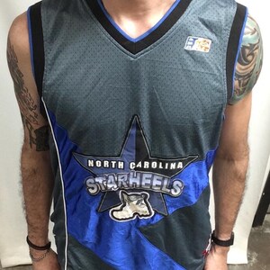 Puede incluir: Una camiseta de baloncesto gris y azul de North Carolina Starheels. La camiseta presenta un gran gr&aacute;fico de estrella con el nombre y el logotipo del equipo. La camiseta tiene cuello en V y ribete negro.