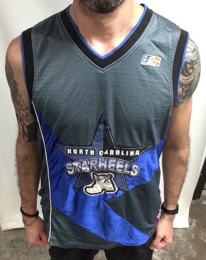 Puede incluir: Una camiseta de baloncesto gris y azul de North Carolina Starheels. La camiseta presenta un gran gr&aacute;fico de estrella con el nombre y el logotipo del equipo. La camiseta tiene cuello en V y ribete negro.