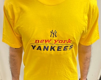 May include: Yellow t-shirt with the New York Yankees logo and text. The logo is navy blue, the text "new york" is red, and "Yankees" is navy blue. The shirt has a crew neck and short sleeves. A classic sports-themed garment.