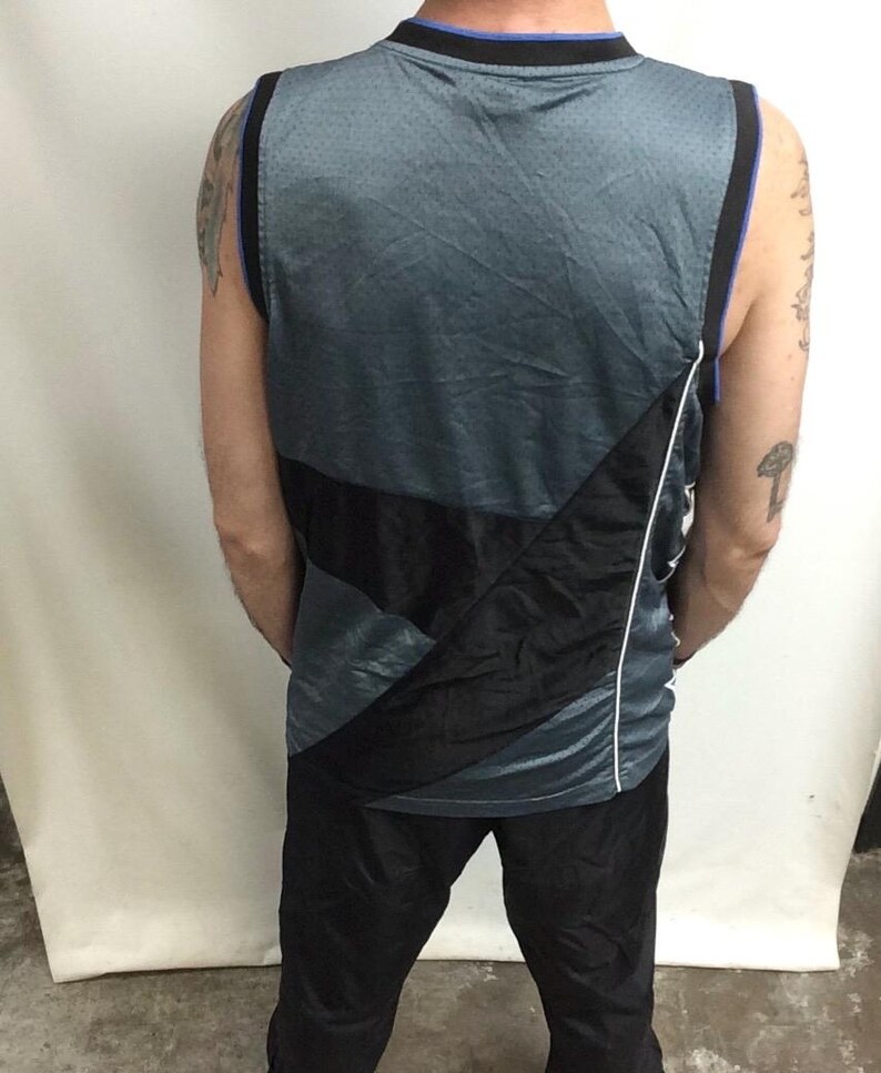 Puede incluir: Camiseta de baloncesto gris y negra con ribete azul alrededor del cuello y las sisas. La camiseta tiene un dise&ntilde;o diagonal negro en la parte delantera y una raya blanca en el lateral. La camiseta se combina con pantalones negros.