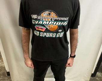 May include: Black t-shirt with a basketball graphic, the text reads "NBA Finals Western Conference Champions" and "Go Spurs Go!". The graphic has orange, teal, and white details. The shirt is worn with black trousers.
