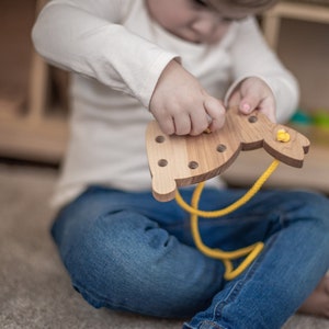 May include: A child is threading a yellow cord through holes in a wooden bunny toy. The bunny is light brown and has a yellow nose and ears.