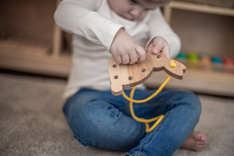 May include: A child is threading a yellow cord through holes in a wooden bunny toy. The bunny is light brown and has a yellow nose and ears.