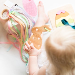May include: A child plays with a wooden bunny toy, a rainbow ribbon toy, and a wooden egg puzzle. The child is wearing a white dress and has blonde hair.