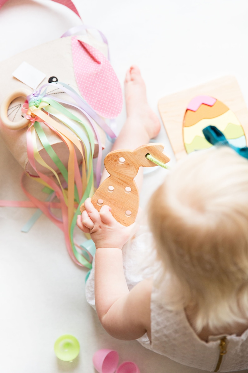 May include: A child plays with a wooden bunny toy, a rainbow ribbon toy, and a wooden egg puzzle. The child is wearing a white dress and has blonde hair.