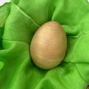 May include: A wooden egg with a light brown finish sits nestled in a bright green fabric. The egg has a smooth, slightly textured surface.