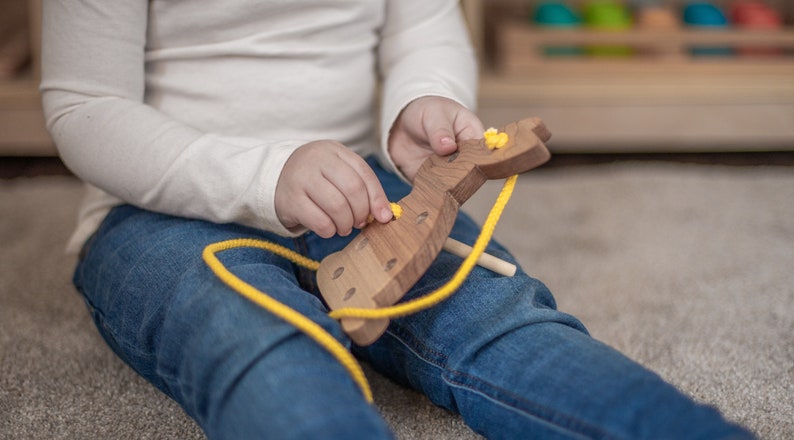 May include: A child is playing with a wooden toy animal. The toy is brown and has yellow buttons. The child is threading a yellow string through the holes in the toy.